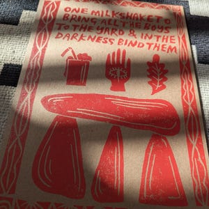 K&ouml;nnte beinhalten: Ein roter und wei&szlig;er Druck auf braunem Papier mit dem Text "ONE MILKSHAKETO BRING ALL THE BOYS TO THE YARD & IN THE DARKNESS BIND THEM". Der Druck zeigt einen roten Milchshake, eine rote Hand mit einem Auge und ein rotes Eichenblatt.
