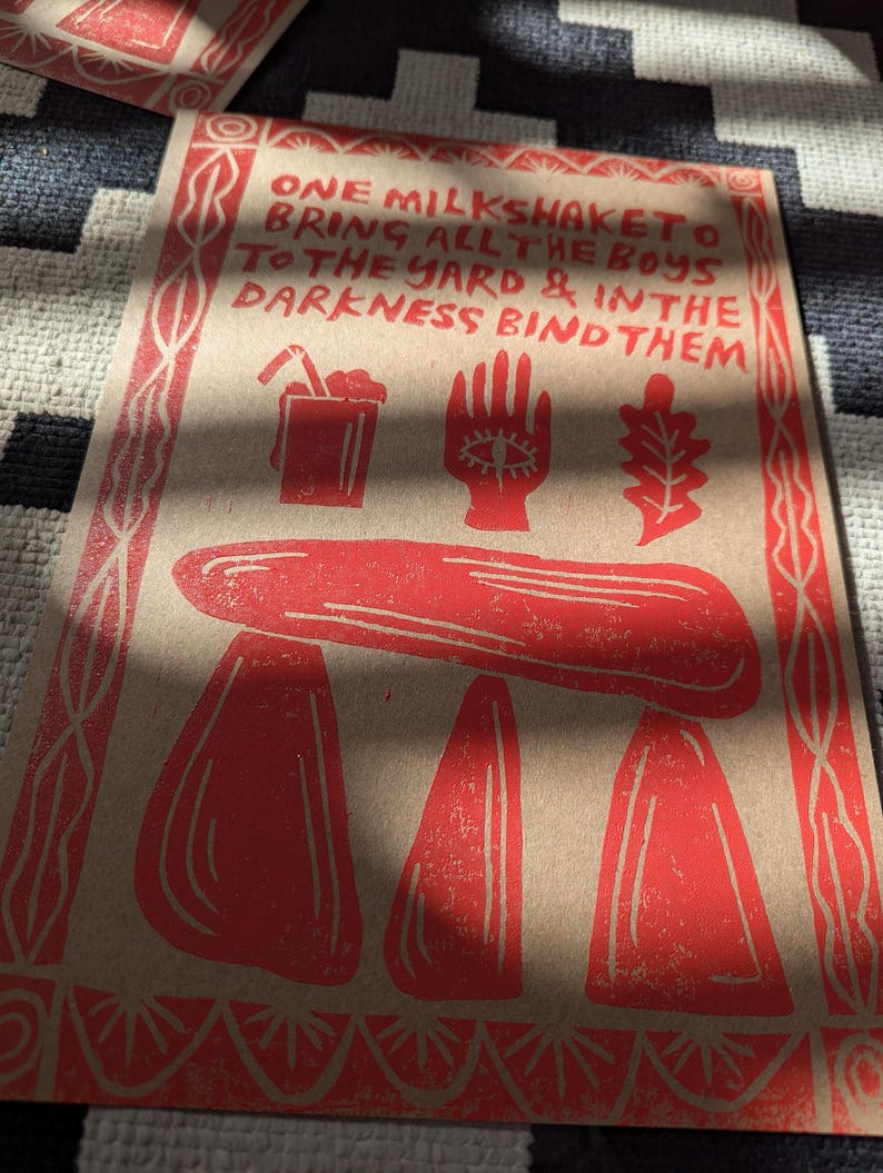 K&ouml;nnte beinhalten: Ein roter und wei&szlig;er Druck auf braunem Papier mit dem Text "ONE MILKSHAKETO BRING ALL THE BOYS TO THE YARD & IN THE DARKNESS BIND THEM". Der Druck zeigt einen roten Milchshake, eine rote Hand mit einem Auge und ein rotes Eichenblatt.