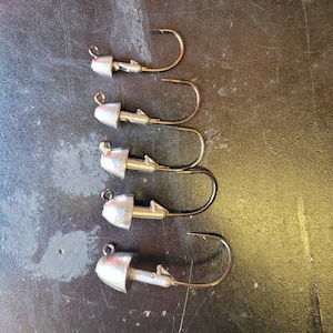 May include: Five silver fishing jig heads with black hooks. The jig heads are different sizes and have a rounded shape.