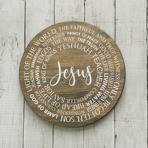 May include: A round wooden sign with the word "Jesus" in the center. The sign is surrounded by a circle of words describing Jesus, such as "Light of the World", "King of Kings", "Wonderful Counselor", "Son", "Lamb of God", "Jehovah", "Emmanuel", "Logos", "The Way", "Yeshuah", "Teacher", "The Faithful and True Witness", "Good Shepherd", "The Life Living", "Alpha & Omega", "Water", "Bread of Life", "Comforter", "Savior", and "Shepherd".
