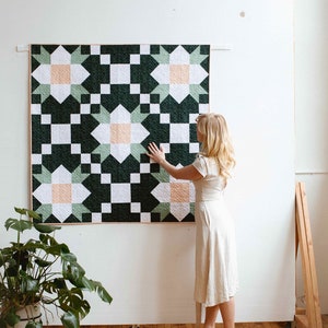 Könnte beinhalten: Eine grüne, weiße und pfirsichfarbene Steppdecke mit einem Sternmuster hängt an einer weißen Wand. Eine Person steht rechts von der Steppdecke und berührt sie mit der Hand.