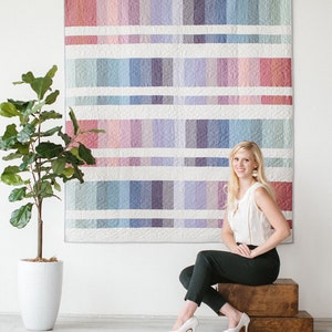 May include: A woman sits in front of a large quilt with a colorful geometric pattern. The quilt is made up of squares of different shades of pink, purple, blue, and green. The woman is wearing a white sleeveless top, black pants, and white heels.