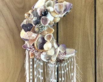 May include: A crescent moon-shaped wall hanging crafted from various seashells in shades of white, tan, purple, and brown. The moon is adorned with dangling white fringe. The background is a wooden wall.