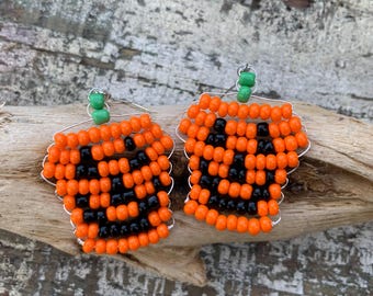 May include: A pair of handmade earrings crafted from orange and black seed beads arranged to form a jack-o'-lantern pumpkin face. Each earring features a small green bead at the top, representing the stem, and is attached to a silver-toned wire hook.