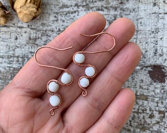 Puede incluir: Un par de pendientes de alambre de cobre hechos a mano con un diseño ondulado. Cada pendiente presenta tres cuentas blancas y redondas. Los pendientes se exhiben sobre una superficie de madera clara y desgastada.