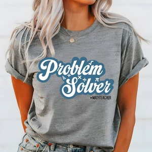 May include: A grey t-shirt with rolled sleeves displaying the text "Problem Solver" in a white and blue design. The shirt includes mathematical symbols and the text "-MATHTEACHER" below. A casual, everyday tee.