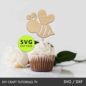 May include: Wooden bee cupcake topper with a light brown color. The bee has a striped body and wings. The topper is on top of a cupcake with white frosting and gold sprinkles. The text "SVG CUT FILES" is on a green circle.