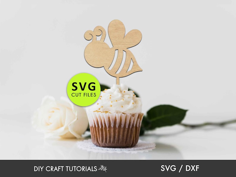 May include: Wooden bee cupcake topper with a light brown color. The bee has a striped body and wings. The topper is on top of a cupcake with white frosting and gold sprinkles. The text "SVG CUT FILES" is on a green circle.