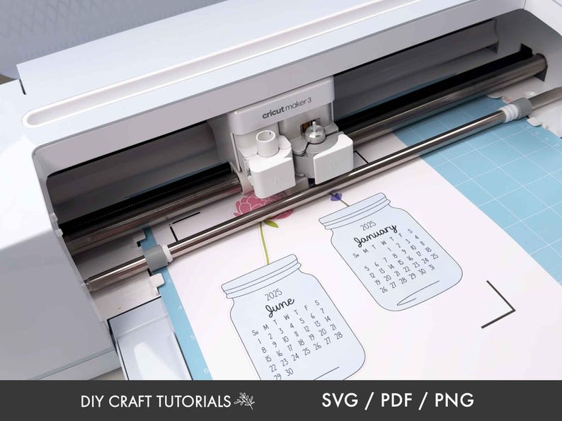 以下が含まれることがあります： 白い Cricut Maker 3 カッティングマシンが、2 つのカレンダーのデザインを切り抜いています。カレンダーはメイソンジャーの形をしており、1 月と 6 月の月が表示されています。各カレンダーには「2025」という文字が印刷されています。