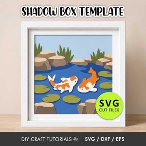 May include: A white square frame containing a layered paper shadow box art piece depicting a blue pond with two orange and white koi fish, green lily pads, and brown rocks. The text SHADOW BOX TEMPLATE and SVG CUT FILES appear on the image.