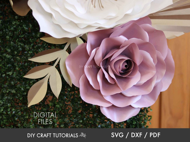 May include: A large, dusty rose paper flower with multiple layers of petals. The flower is surrounded by greenery and a smaller white paper flower.
