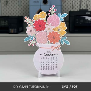 K&ouml;nnte beinhalten: Papierkalender f&uuml;r 2026 in Vasenform mit bunten Papierblumen. Der Kalender zeigt den Monat Januar auf Spanisch, mit einer Schleife und einem St&auml;nder. Der Text "DIY CRAFT TUTORIALS" und "SVG / PDF" befinden sich unten.