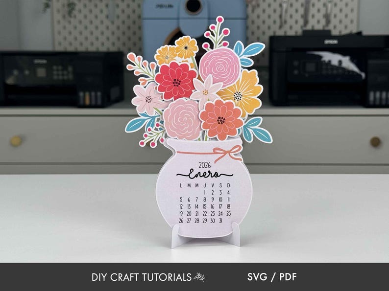 K&ouml;nnte beinhalten: Papierkalender f&uuml;r 2026 in Vasenform mit bunten Papierblumen. Der Kalender zeigt den Monat Januar auf Spanisch, mit einer Schleife und einem St&auml;nder. Der Text "DIY CRAFT TUTORIALS" und "SVG / PDF" befinden sich unten.