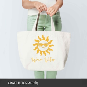 May include: Cream-colored tote bag with a yellow sun wearing sunglasses and the words "Warm Vibes" printed in a script font. The bag has brown handles and is held by a person.