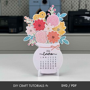 K&ouml;nnte beinhalten: Papierkalender f&uuml;r 2026 in Vasenform mit bunten Papierblumen. Der Kalender zeigt den Monat Januar auf Spanisch, mit einer Schleife und einem St&auml;nder. Der Text "DIY CRAFT TUTORIALS" und "SVG / PDF" befinden sich unten.