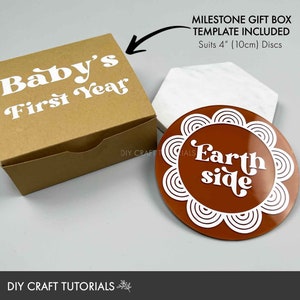May include: A brown cardboard box with the text "Baby's First Year" in white. A brown and white round coaster with the text "Earth side" in white. The box and coaster are part of a milestone gift box set for a baby's first year.