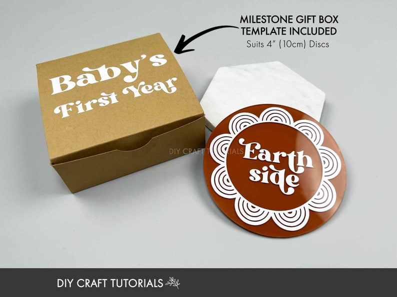 May include: A brown cardboard box with the text "Baby's First Year" in white. A brown and white round coaster with the text "Earth side" in white. The box and coaster are part of a milestone gift box set for a baby's first year.
