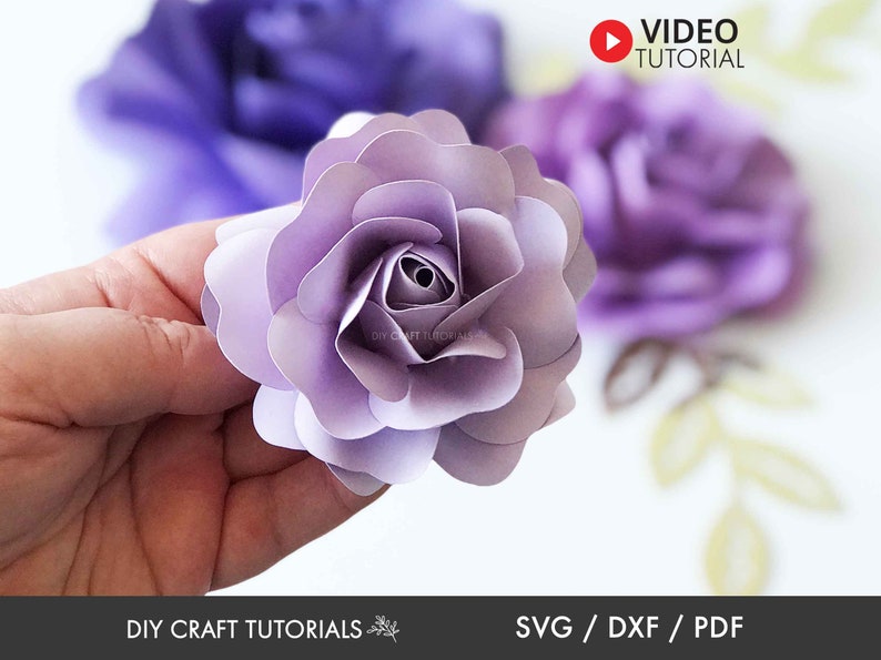 Puede incluir: Primer plano de una rosa de papel de color lila claro. La rosa est&aacute; hecha de varias capas de papel y tiene un aspecto delicado y en capas. La rosa se sostiene en una mano, y el fondo est&aacute; desenfocado.