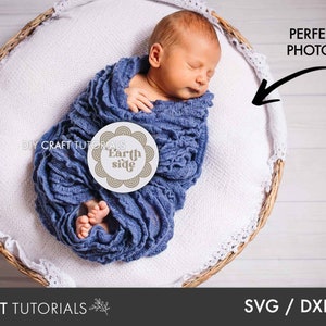 May include: A newborn baby wrapped in a blue blanket is lying in a wicker basket. The baby is holding a round white sign with the text "Earth side" in brown lettering. The sign is decorated with a brown circle pattern.
