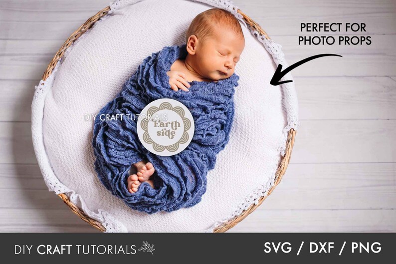 May include: A newborn baby wrapped in a blue blanket is lying in a wicker basket. The baby is holding a round white sign with the text "Earth side" in brown lettering. The sign is decorated with a brown circle pattern.