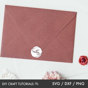 May include: A maroon envelope with a white round sticker that says "Wedding" in a cursive font. The envelope is on a white surface with a small red flower.