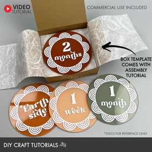 May include: Four round, decorative discs for a baby milestone photo prop. The discs are made of wood and have a white border with a decorative pattern. The discs feature the following text: "Earth side", "1 week", "1 month", and "2 months".