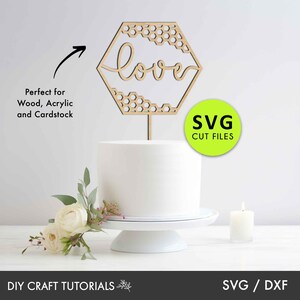 May include: A wooden hexagon cake topper with a honeycomb pattern and the word "love" in a cursive font. The topper is on a white cake stand with a white cake.