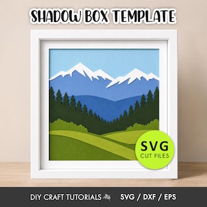 May include: A square white frame containing a layered paper shadow box art piece depicting a mountain landscape with snow-capped peaks, a dark green pine forest, and rolling light green hills. The text SHADOW BOX TEMPLATE and SVG CUT FILES appear.