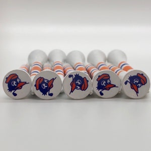 May include: Eight white golf tees with orange and blue stripes and a cartoon bird design. The bird is wearing a hat and holding a golf club.