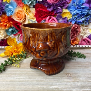 May include: A brown ceramic planter with a textured glaze and a face sculpted into the side. The planter has a wide base and a rounded top.