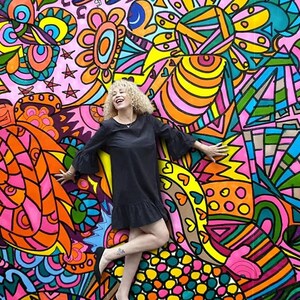 May include: A woman with curly blonde hair is standing in front of a colorful, abstract mural. The mural is painted in bright colors, including pink, yellow, blue, green, and orange. The woman is wearing a black dress and is smiling.