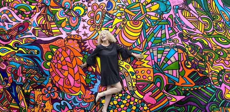 May include: A woman with curly blonde hair is standing in front of a colorful, abstract mural. The mural is painted in bright colors, including pink, yellow, blue, green, and orange. The woman is wearing a black dress and is smiling.
