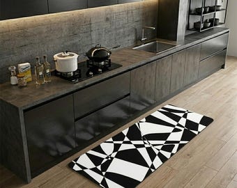 May include: Modern kitchen with black cabinets, a stainless steel sink, and a black and white geometric patterned floor mat. A stovetop with a pot and pan is visible. The kitchen has a minimalist design.