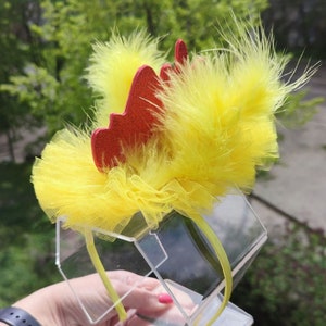 Puede incluir: Diadema amarilla y roja con plumas y tul. La diadema está en un soporte de plástico transparente.