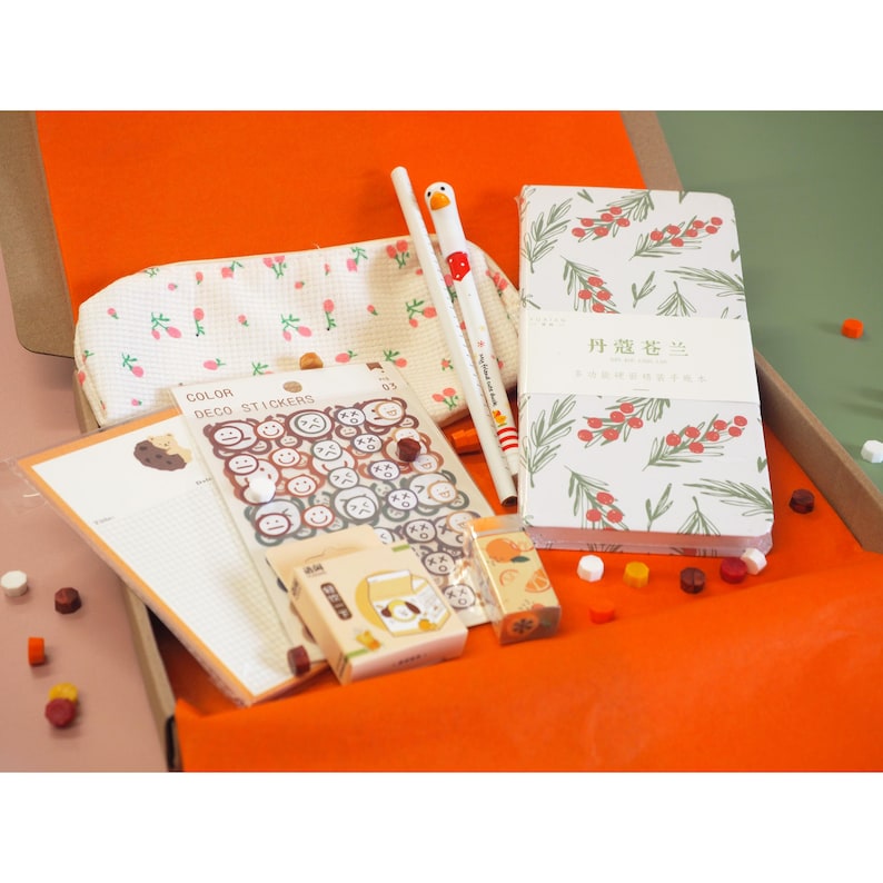 May include: An open orange box filled with stationery items. Includes a pencil case, notebooks with a floral design, stickers, pens, and small boxes. The items have a pastel color palette with a focus on white, green, and orange.