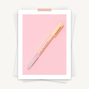 May include: A light peach-colored pen with a matching cap and clip, lying diagonally on a pink background. The pen has the words "BLUE PEN..." printed on its side. The pen is set on a white paper background.