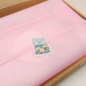 May include: A small, colorful stamp featuring a landscape scene with trees and water, placed on a sheet of pink tissue paper inside a cardboard box. The stamp has a white border and a detailed illustration.