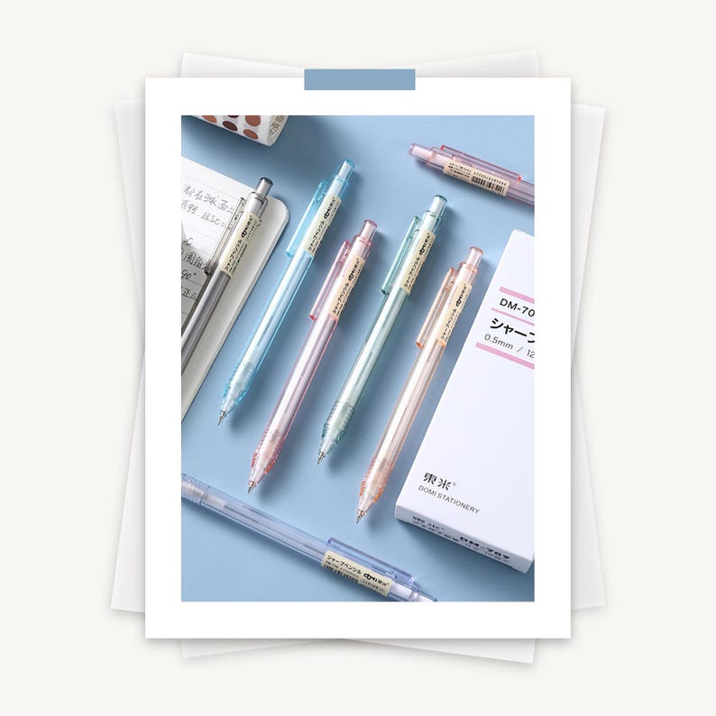 K&ouml;nnte beinhalten: F&uuml;nf durchsichtige mechanische Bleistifte mit verschiedenen Farben: blau, rosa und hellblau. Die Stifte haben einen Durchmesser von 0,5 mm und einen transparenten Radiergummi am Ende. Die Stifte sind in einer Reihe auf einer hellblauen Oberfl&auml;che angeordnet. Der Text auf der Schachtel lautet "DM-70 シャープ 0.5 mm / 12" und "DOMI STATIONERY".