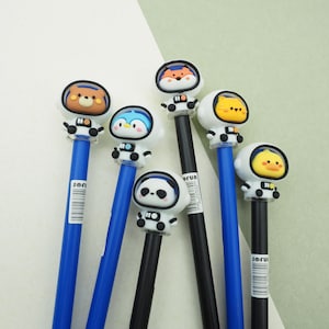 May include: A collection of pens featuring cartoon animal astronaut toppers. The pens have blue and black barrels, with animal faces including a bear, penguin, fox, cat, panda, and duck. The pens are arranged on a light green and white background.