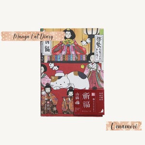 May include: A red and gold Japanese-style diary with a cat illustration and the text "Manga Cat Diary" and "啓蟄" on the cover. The diary is accompanied by a small red and gold charm with the text "祈福" and "山猫" on it.