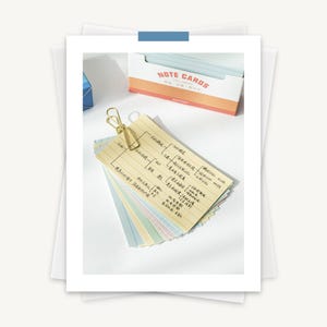 May include: A stack of yellow note cards with lined paper and a gold paper clip. The cards have handwritten text on them, including the words "remarkable" and "time". The cards are sitting on a white surface.