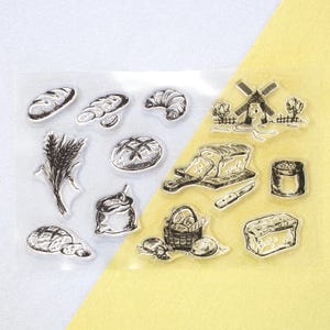 May include: Clear stamp set featuring various bread-related illustrations. Includes images of loaves, a croissant, wheat, a windmill, and a bread basket. The stamps are in a sketched style, perfect for crafting and scrapbooking projects.