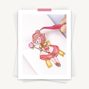 May include: A colorful sticker of a cartoon girl in a pink dress holding a pink flower. The sticker is being held by pink tweezers. The girl has brown hair and a pink bow. The sticker is on a white background.