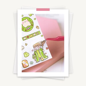 May include: A sheet of colorful stickers featuring cartoon illustrations of a bear, bread, and a character in a green cloak. The stickers are being held by pink tweezers. The background includes a pink notebook and white paper.