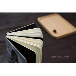 May include: A black hard cover notebook with a colorful illustration of a fish on the front. The notebook is open to reveal different types of paper, including lined, grid, and blank. The notebook is 25mm thick.