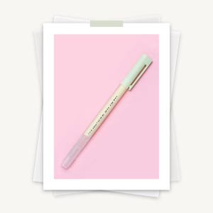 May include: A light green pen with a matching cap, lying diagonally on a pink background. The pen's barrel is a pale pink, and the text "...GLUE PEN..." is printed on the side. The pen is a stationery item.
