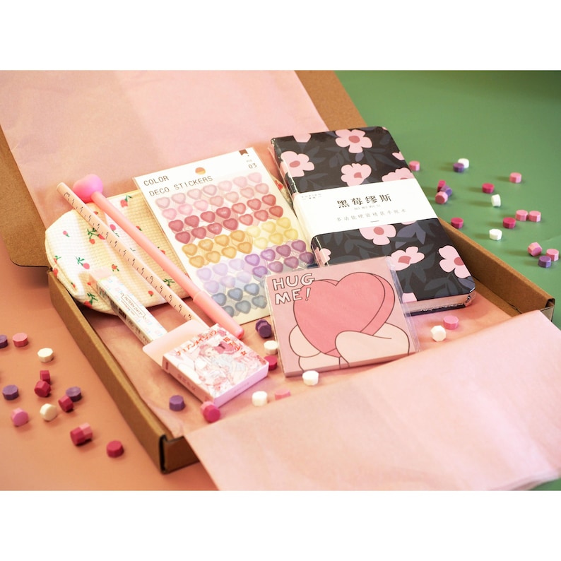 May include: A gift box filled with stationery items, including a notebook with a floral design, heart-shaped stickers, a pink pen, and a notepad with a heart graphic that says "HUG ME!". The box is lined with pink paper.
