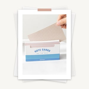 May include: A blue and white box of note cards with the text "NOTE CARDS" in a white circle. The box is open and a hand is pulling out a light brown note card with a grid pattern.