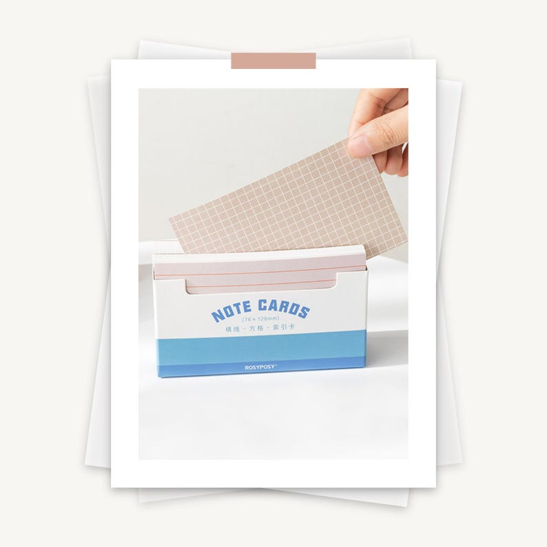 May include: A blue and white box of note cards with the text "NOTE CARDS" in a white circle. The box is open and a hand is pulling out a light brown note card with a grid pattern.