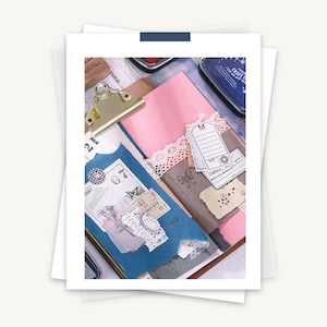 May include: A close-up of a scrapbook page with various decorative elements, including paper scraps, stickers, stamps, and tags. The page is decorated with a pink and blue color scheme and features a gold binder clip.